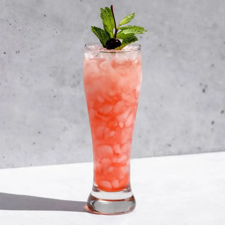 A tall pilsner glass holds a bright red drink over crushed ice, garnished with mint and a cherry.