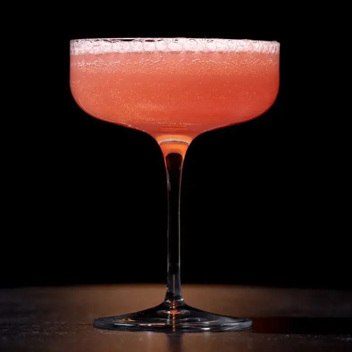 A dark pink sparkling cocktail in a wide, angular coupe pictured against a black background