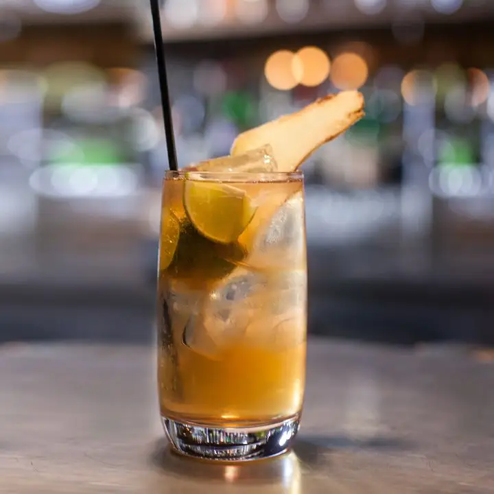 Brazilian Buck cocktail in a highball glass, garnished with lime wedges and ginger peel, served with a black straw