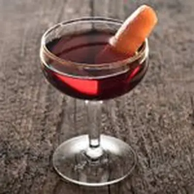 A short, unadorned coupe stands on a wooden surface. The glass holds a crimson cocktail and a thin slice of orange.
