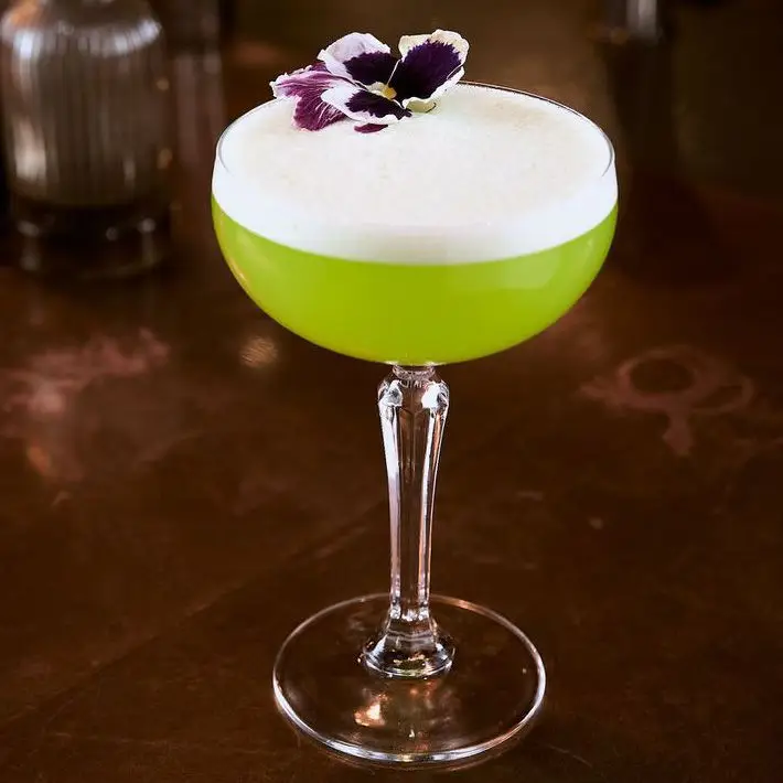 Bright green Yuzu Midori Highball in coupe glass on bar top, with thick white foam and flower garnish