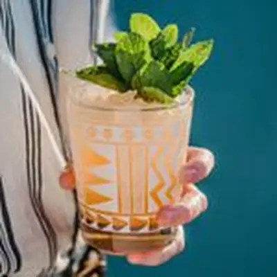hand holds the Cheeky Peach cocktail, in a rocks glass with crushed ice and garnished with mint leaves