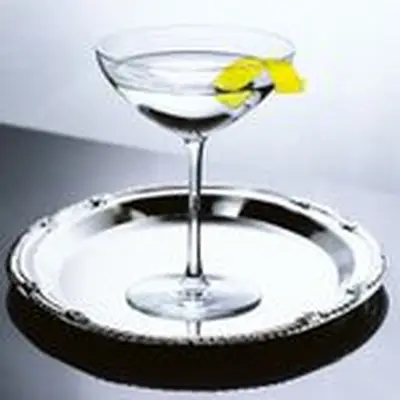 A swooping martini glass sits on a silver platter. Only the bright yellow lemon peel draped over the lip of the glass gives the otherwise crystalline and silver image any color