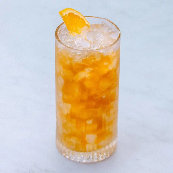 A Collins glass filled with crushed ice rests on a marble surface. The drink within is golden brown with sherry, garnished with an orange peel.