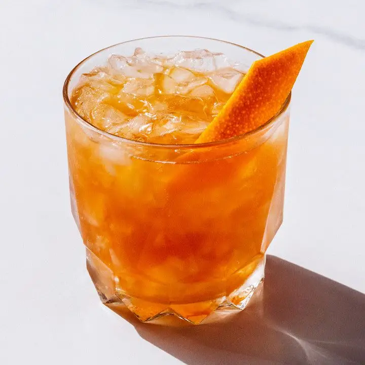 A rocks glass sits on a marble surface. It’s filled with crushed ice and an orange-colored cocktail, and garnished with an orange peel.