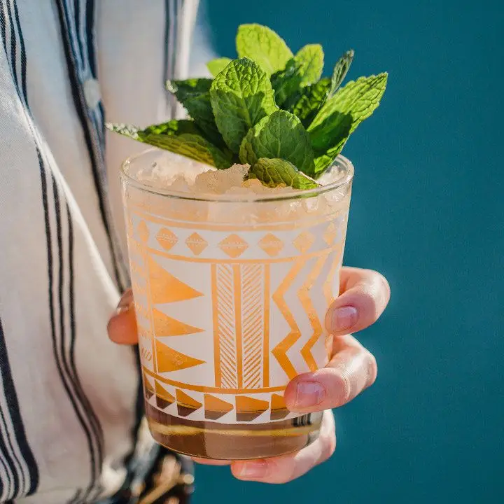 hand holds the Cheeky Peach cocktail, in a rocks glass with crushed ice and garnished with mint leaves
