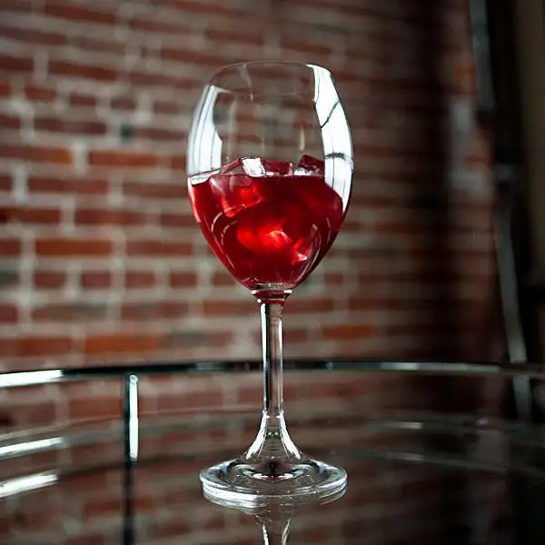 A wine glass with a thick stem and delicate bowl holds a bright crimson drink over a few ice cubes. A brick wall is out of focus in the backdrop