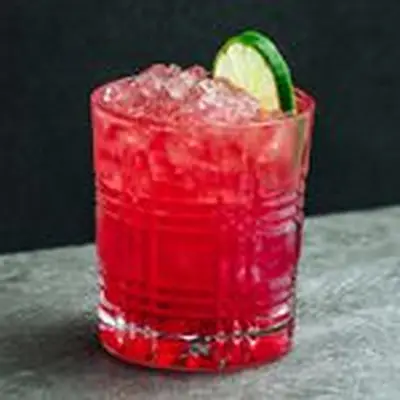 A detailed rocks glass rests on a gray stone surface against a black backdrop. The glass is filled with a vivid red drink over crushed ice and garnished with a lime wheel.