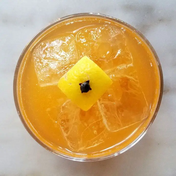 An overhead shot of a bright orange drink over ice, resting on a marble surface. It’s garnished with a lemon peel square pierced in the center by a clove. 