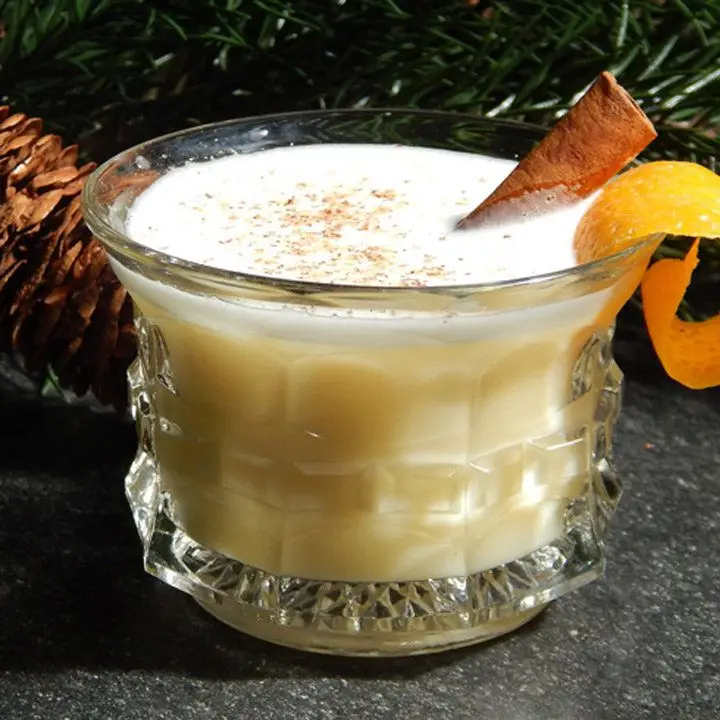 creamy yellow Trick Nog cocktail in ornate punch glass with cinnamon stick and orange peel garnishes, sitting in front of a pine tree