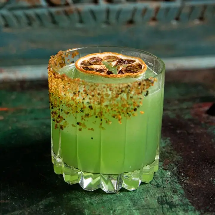 A faceted rocks glass rests on a green bar top. The glass is partially coated in chile salt, and the drink within is a vivid green. It’s garnished with a dehydrated blood orange wheel. 