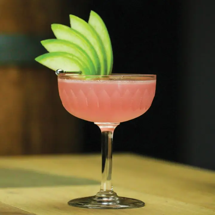 pink-hued rosemary & rhubarb cocktail in a coupe, garnished with an apple fan
