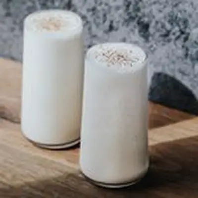 Two tall glasses sit on a wooden surface, a blue and white speckled wall behind them. The glasses are filled to the brim with a creamy white drink and are dusted with nutmeg.
