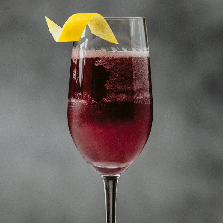 The bowl of a champagne flute is shown in a close-up shot. It’s filled with a bubbly violet drink and garnished with a small, curled lemon twist. The background is gray and fuzzy.