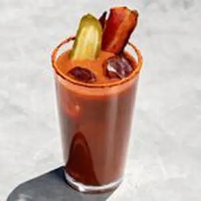 A Bloody Mary in a tall, narrow pint glass rests on marble. The drink is garnished with a pickle skewer and bacon slice, and the glass is rimmed with dusty red spices.