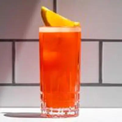 An etched Collins glass containing a frothy orange-red cocktail, garnished with an orange wedge and set against a white subway-tiled background