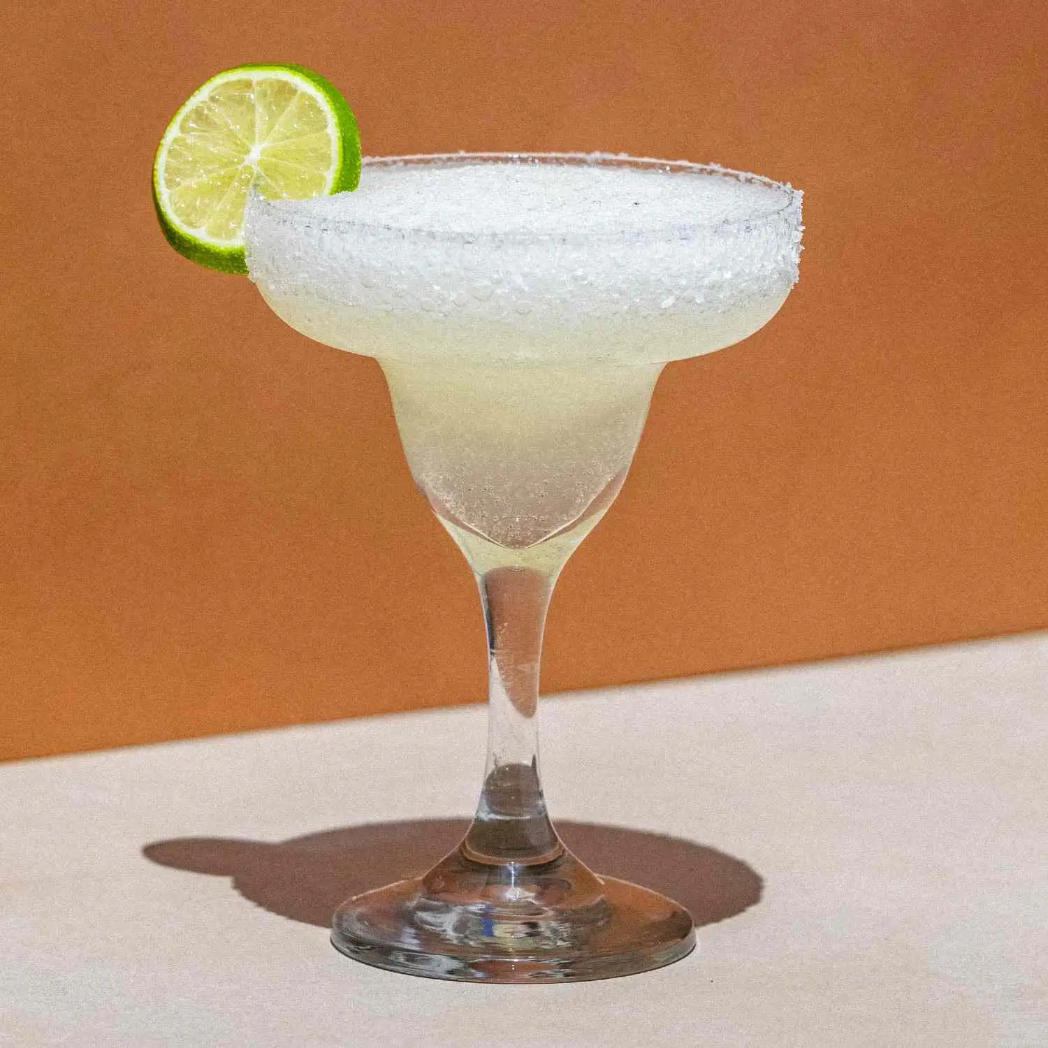 Frozen Margarita in curved margarita glass with salt rim and lime wheel, on orange background