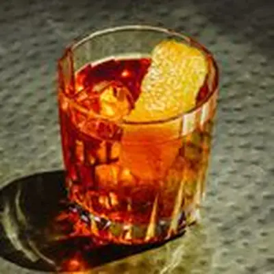 A rocks glass on a dimpled dark surface holds an amber-hued drink over ice cubes. It’s garnished with an orange peel.