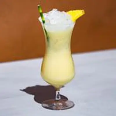 Painkiller cocktail, pale yellow, in a curved hurricane glass over crushed ice, with a bamboo straw and pineapple wedge garnish