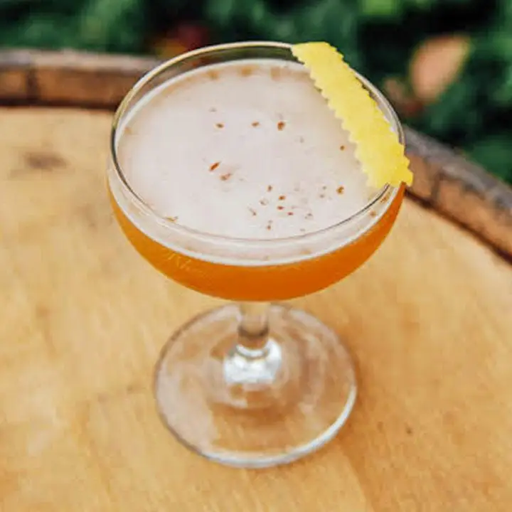A cocktail coupe sits on a wooden barrel, and is filled with an orange drink. A pinked lemon peel rests on the mouth of the glass. 