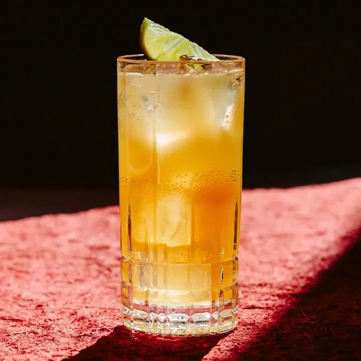 A faceted Collins glass holds a red-orange drink garnished with a lime wedge. It sits on a red stone surface with dramatic lighting.