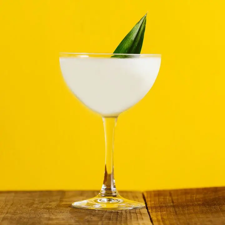 A creamy white cocktail in a coupe glass garnished with a pineapple leaf, set on a wood surface against a bright yellow background