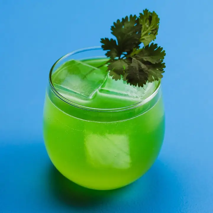 A round rocks glass is set against a solid blue backdrop. The drink within is vivid green, with a few ice cubs and a sprig of cilantro.