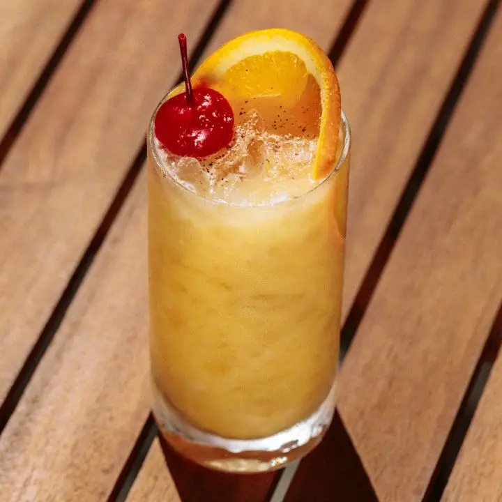A Collins glass rests on wooden slats. The drink within is cloudy and yellow over crushed ice, garnished with a cherry and orange wheel.