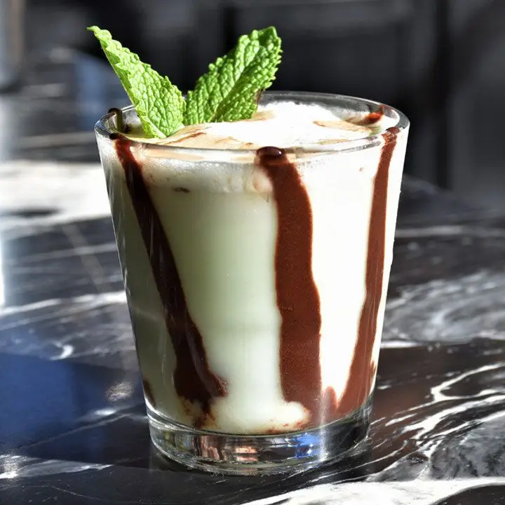 A shot glass sits on a black marble countertop; it is filled with a green, creamy elixir and lines of chocolate streak it like an inverse of the counter top. It is garnished with two small mint leaves.