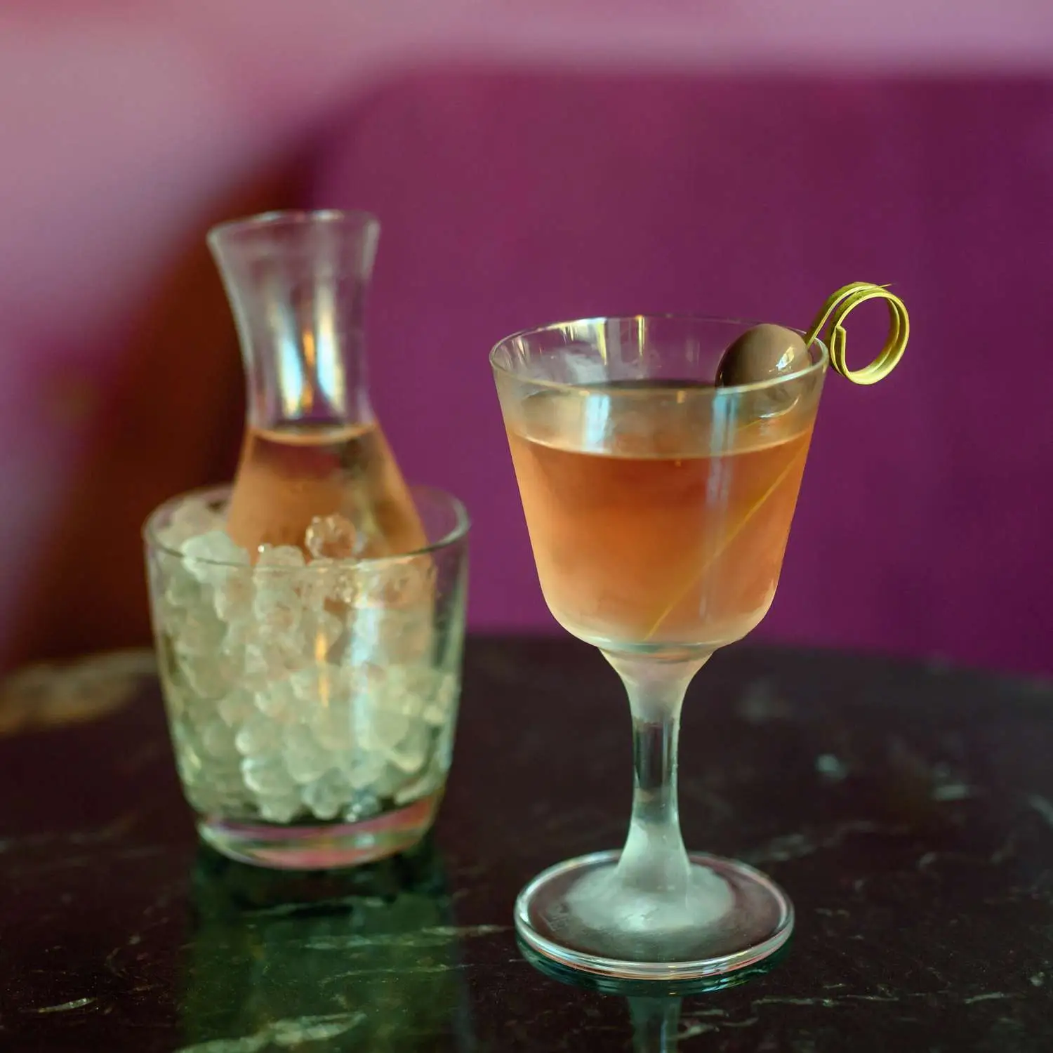 Pink-hued Miladys Martini in Nick and Nora glass with purple olive on a toothpick, with sidecar in background, shot on black marble table with pink background
