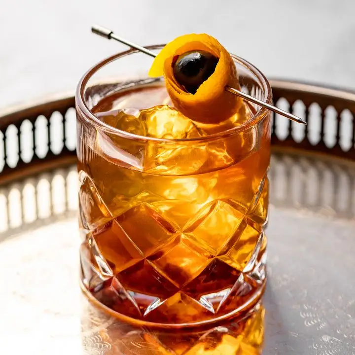 Brown Butter Old Fashioned cocktail in a crystal-cut rocks glass with a skewered cherry rolled in an orange peel, served on a round metal tray