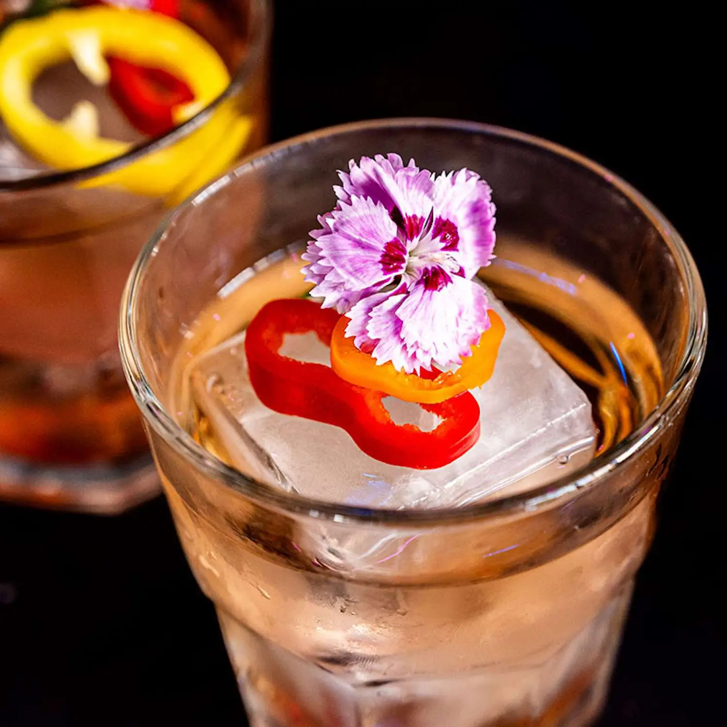 A Little Chili Punch cocktail in a rocks glass with a single large ice cube, and flower and pepper garnish