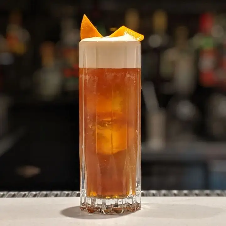 A tall, chunky collins glass sits on a bar top, and holds a dark amber liquid and a few large ice cubes. On top, a curved orange zest floats on a thick layer of foam. 