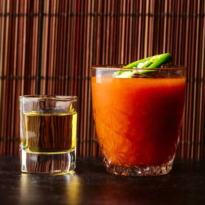 Two glasses rest on a black surface in front of a bamboo mat. On the left is a squat shot glass of tequila. On the right is a larger glass of vivid orange Sangrita, garnished with a jalapeño.