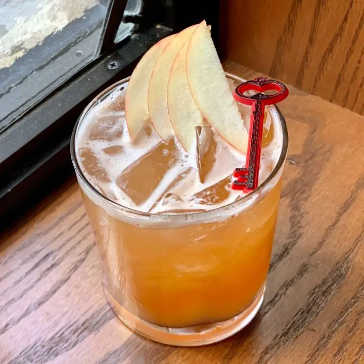 A short rocks glass sits in the corner of a wooden windowsill, a window just out of frame behind it. The glass is filled with ice and a red-orange drink, and is garnished with four thin slices of apple and a plastic red and black key. 