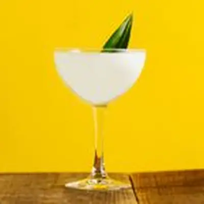 A creamy white cocktail in a coupe glass garnished with a pineapple leaf, set on a wood surface against a bright yellow background