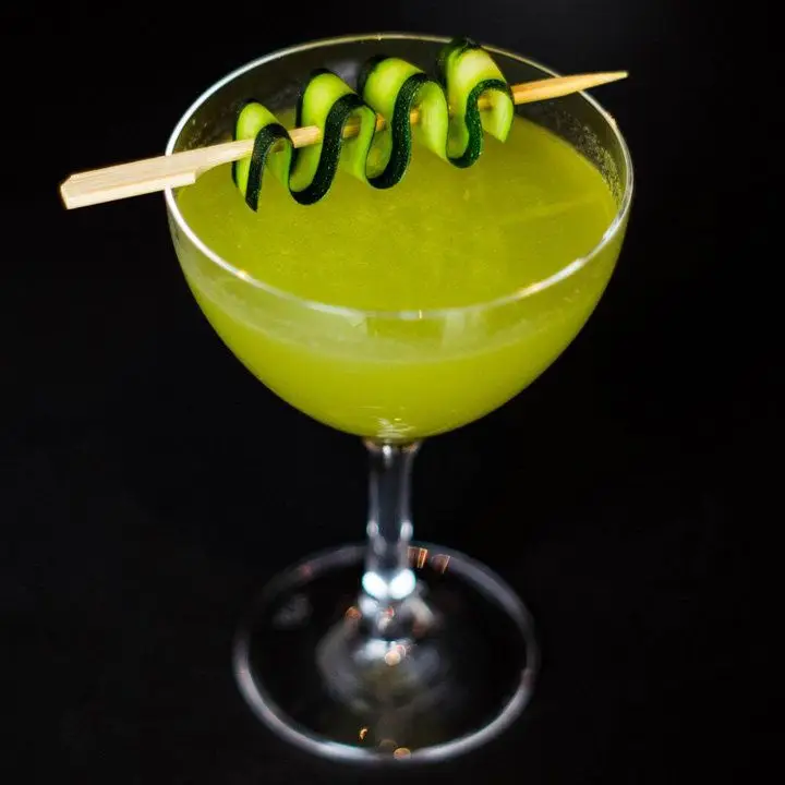 bright-green Fields of Travel cocktail in a coupe with a skewered zucchini ribbon balanced on the glass