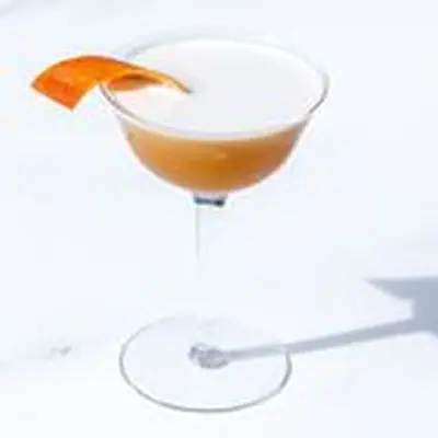 Brown Derby cocktail in high-stemmed coupe glass, with a head of foam and orange twist, on a light colored background