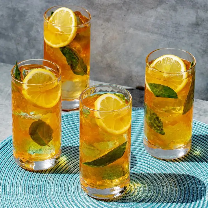 Four Collins glasses sit on a blue mat on concrete. Each glass is filled with iced tea over ice with a large lemon wheel and mint leaves.