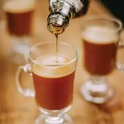 Maple syrup is poured into one of three frothy Hot Butter(nut) Rum cocktails, each served in a toddy glass with handle