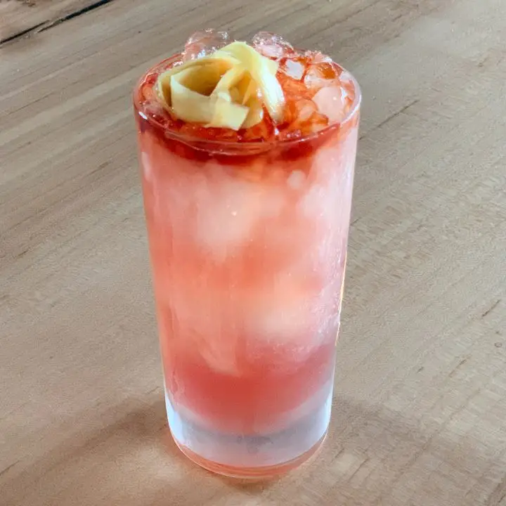 A pinkish-red cocktail in a Collins glass topped with Angostura and fresh ginger
