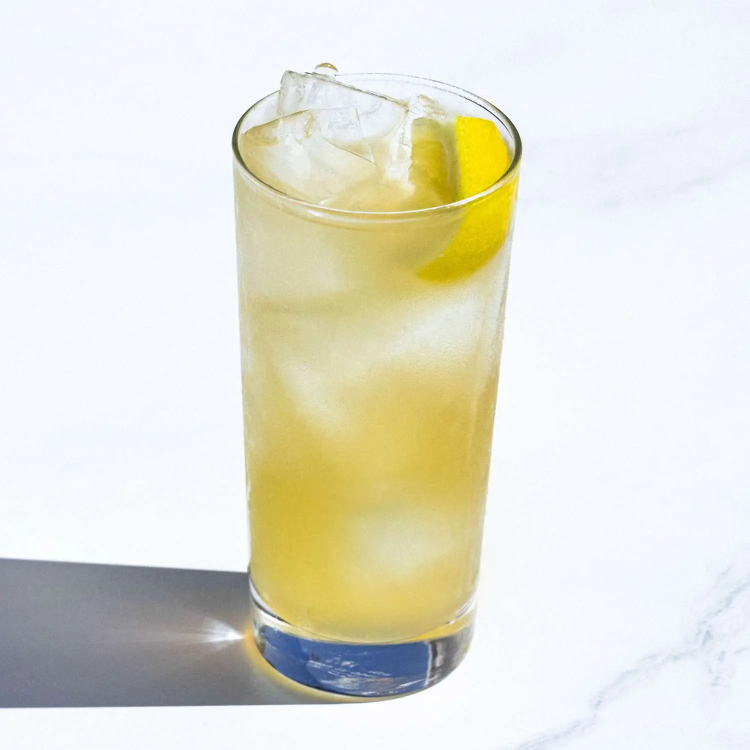 Horsefeather cocktail, light yellow, in Collins glass filled with ice and lemon wedge garnish