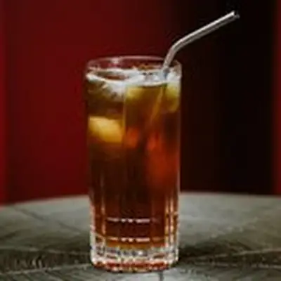 A Collins glass with a metal straw rests on a ringed mat. The drink within has a few ice cubes and is dark brown.