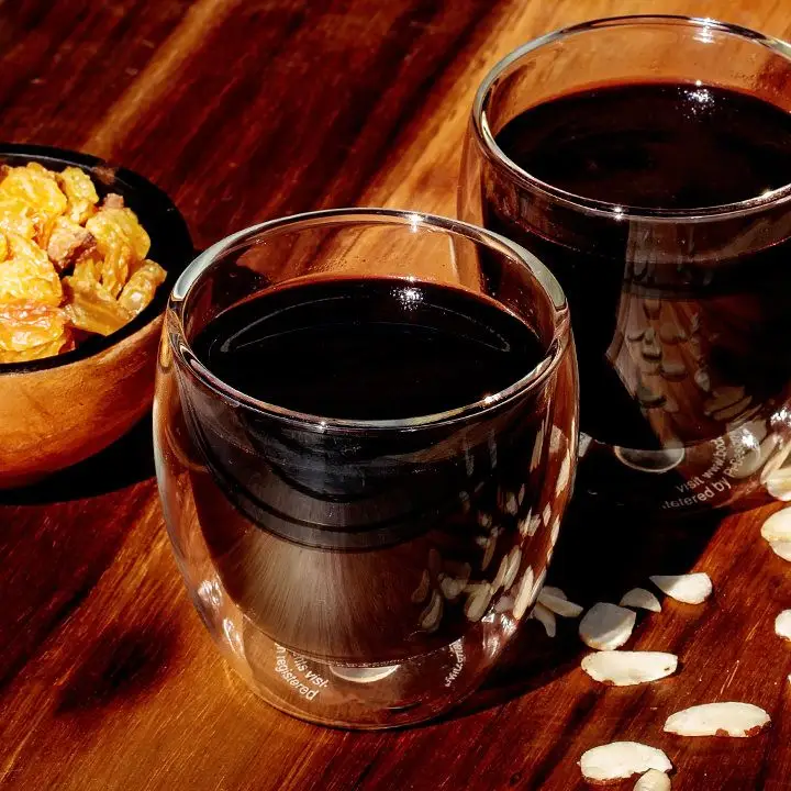 A dark-red Glögg cocktail in a stemless wine glass on a table with scattered almond slices and a bowl of dried fruit.