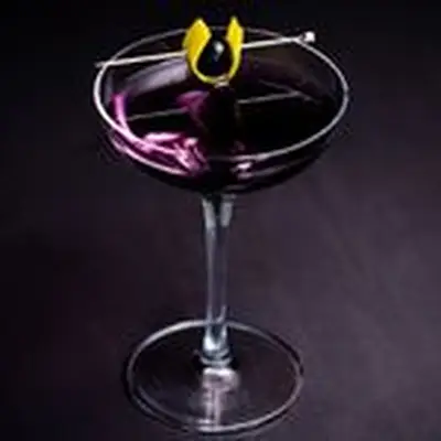 An elegant coupe glass holds a deep violet drink, garnished with a cherry wrapped in a lemon peel, pierce by a pick. The glass sits on a dark gray surface.