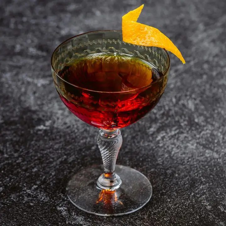 An elaborate cocktail glass with a detailed stem holds a vivid red Old Fashioned. The fancy glass is garnished with a twist of orange zest and sits on a black and gray surface