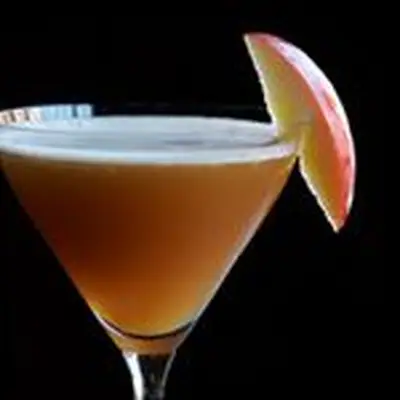 On a solid black background, a cocktail glass is filled with a dark brown drink, a thin layer of white foam resting on top. It’s garnished with a sliver of apple.