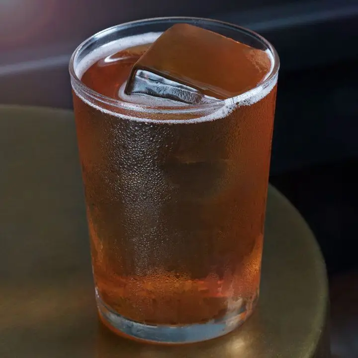 A short Collins glass rests on a bronze stool. The glass is filled with a few large, clear ice cubes and a dark brown highball. 