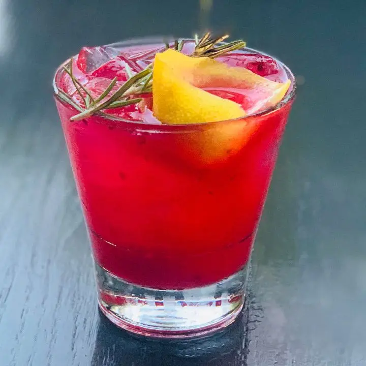 red-colored Delete That Text cocktail in a rocks glass, garnished with a rosemary sprig and curled lemon peel