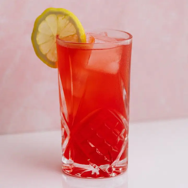 A tall Collins glass is filled with a bright red drink and a few ice cubes. It’s garnished with a lemon wheel. The backdrop is pale pink.